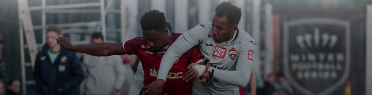 На изображении футболисты в динамичном моменте матча: игрок в форме бордового цвета пытается прорваться сквозь защиту игрока в серой форме. На заднем плане видны другие люди и часть спортивной инфраструктуры