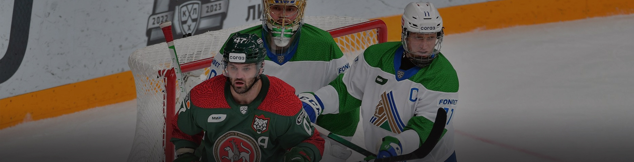 На изображении три хоккеиста в процессе игры: один в зелёно-красной форме с чёрным шлемом и двое в бело-зелёной форме, один из которых вратарь в шлеме и с маской. На заднем плане видна часть хоккейных бортиков и логотип