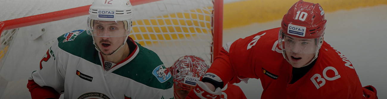 На изображении два хоккеиста в экипировке играют на льду возле ворот. Один игрок в белой форме с зелёным, другой в красной форме. Оба активно участвуют в игровом моменте