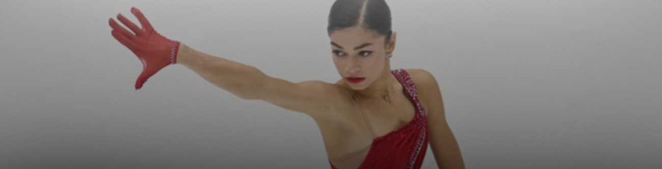 На изображении фигуристка в красном костюме и перчатке исполняет элемент программы на льду. Она вытянула руку в сторону, создавая грациозную позу. Фон размыт, акцент на спортсменке