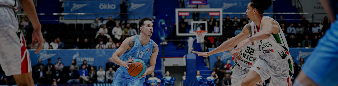 На изображении запечатлен момент баскетбольного матча. Игрок в голубой форме ведет мяч, готовясь сделать передачу или бросок, в то время как противники в белой форме защищаются. На заднем плане трибуны с болельщиками