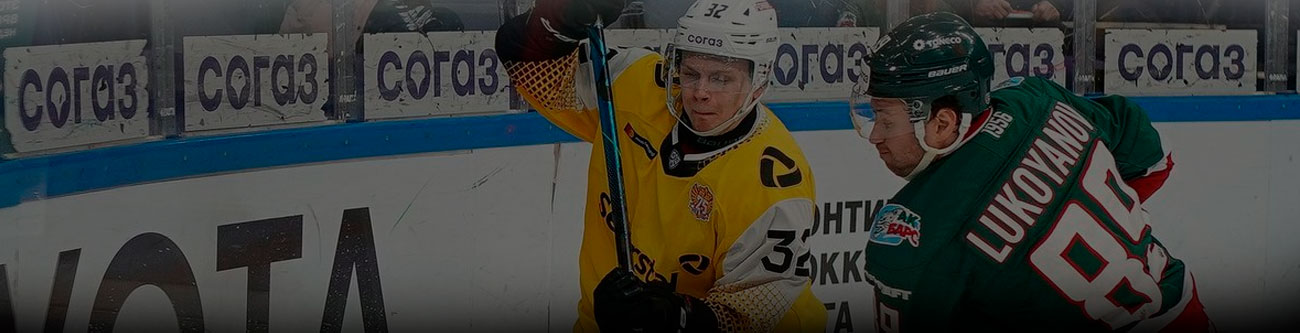 На изображении запечатлен хоккейный матч. Игрок в желтой форме противостоит игроку в зеленой форме около борта. На заднем плане видны рекламные баннеры и ограждение игровой зоны